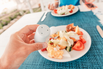 Hand holding salt shaker over plate with salad, potatoes, tomatoes and scrambled eggs during...