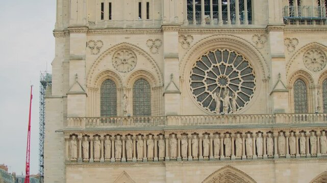 Close upshot of the facade of Notre-Dame Cathedral in Paris, showcasing rose window, rows of stone saints, arched stained-glass windows, Gothic arches, and restoration equipment