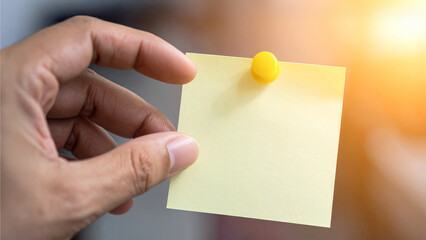 A hand holding a blank yellow sticky note with a push pin, ready for messages