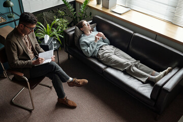 Man lying on sofa talking during therapy session with male therapist sitting nearby writing notes...