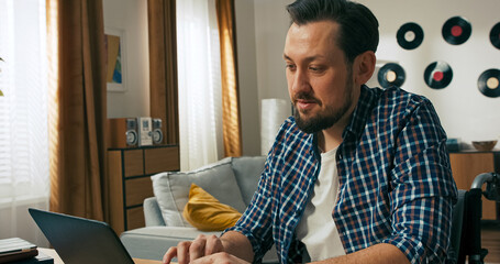 A man with a disability is seated at home, using a laptop for a financial transaction. Holding a...