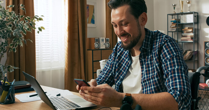 A bearded man with a disability multitasks from home. He writes messages on his computer and watches a video on his phone, exchanging stories with friends.