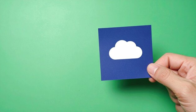 A person's hand holding a blue square paper with a white cloud icon against green background - Powered by Adobe