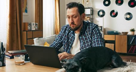 A concentrated young man with a beard, wearing home clothes, sits in a wheelchair and works online from home while his favourite puppy rests nearby, providing comfort and companionship.