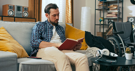 A man with a disability sits alone on the couch reading a book. His expression is sad and thoughtful, spending quiet time at home with his wheelchair beside him in the background.