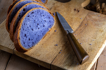 Freshly baked bread with a vibrant purple interior, cooked in blue Thai ancha tea, rests on a rustic wooden cutting board with a slice cut off to reveal its soft, crumbly texture, with copy space