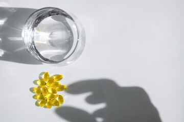 Yellow transparent capsules or pills or vitamins or supplements are near glass of water with hand shadow reaching for them on white background, representing medicine and healthcare