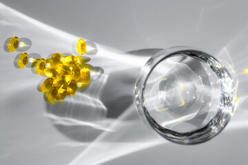 Yellow vitamin D or fish oil capsules are scattered near a glass of water, casting intriguing...