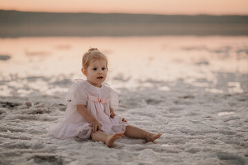 Baby girl in a light dress and two ponytails at sunset on the sea in summer