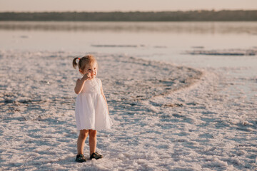 Baby girl in a light dress and two ponytails at sunset on the sea in summer