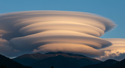 A spectacular multi-layered lenticular cloud formation, resembling a UFO, glowing at sunset over a mountain peak. Rare and breathtaking atmospheric weather phenomenon. A surreal and majestic
