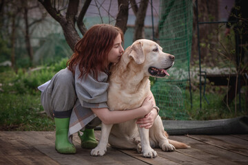A teenage girl in rubber boots and a poncho hugs her Labrador retriever in a quiet garden full of trees and lush grass.