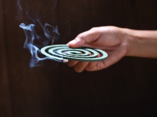 A person's hand holds a circular mosquito coil with the tip burning and smoking.