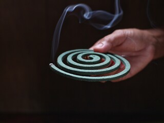 A person's hand holds a circular mosquito coil with the tip burning and smoking.