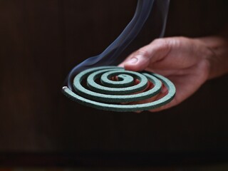 A person's hand holds a circular mosquito coil with the tip burning and smoking.