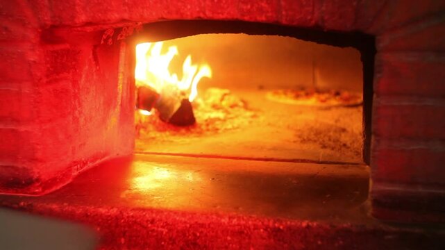 Traditional pizza being baked in a hot wood-fired oven while handled with a wooden pizza peel near the open flame.