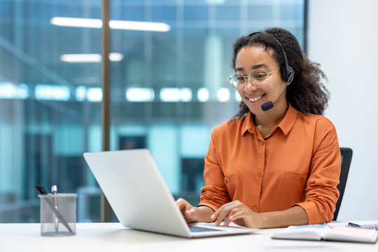 Smiling customer service representative wearing a headset and glasses working on a laptop in a modern office setting. Focus on communication and support.