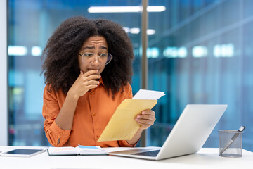 A concerned young woman with glasses examines documents while working at her office desk, looking...
