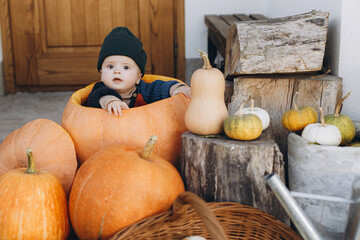 Cute baby boy sitting in big pumpkin at front door. Happy halloween and Thanksgiving. Adorable little child in cozy sweater sitting with pumpkins on porch, autumn decor at home