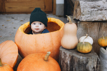 Sweet baby boy playing in big pumpkin at front door with autumn rustic decor. Happy halloween and Thanksgiving. Cute little child in cozy sweater sitting with pumpkins on porch