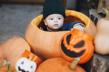 Trick or treat! Sweet happy baby boy in big pumpkin at front door with jack o lantern and autumn decor. Happy halloween. Adorable little child sitting with pumpkin bucket on porch