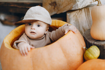 Sweet baby boy playing in big pumpkin at front door with autumn rustic decor. Happy halloween and Thanksgiving. Adorable little child in sitting with pumpkins on porch