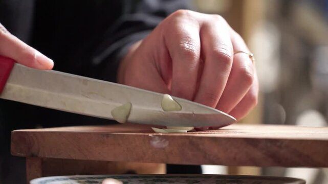 Close-up shot of hands slicing garlic on a wooden cutting board with a kitchen knife. Natural lighting and shallow depth of field emphasize a calm cooking moment.