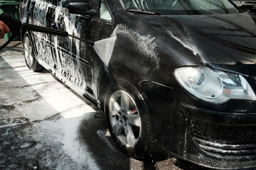 Side view of a black car being pressure washed, with thick foam and water splashing during cleaning.