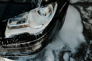 Close-up top view of a car headlight and bumper covered in soap suds, with foam dripping onto the...
