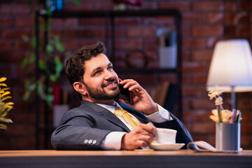 Indian businessman talks on phone while enjoying tea, showing relaxed yet connected office routine