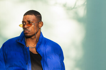 Confident African American male wearing trendy orange sunglasses and warm jacket leaning on wall in...