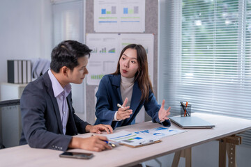 Two people in suits are talking at a desk with a lot of papers and graphs