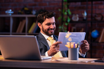 Indian businessman smiles at reports while checking printed financial results at office desk