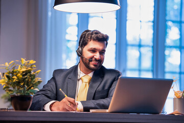 Indian businessman writes notes during online call, showing focus, learning, and remote teamwork