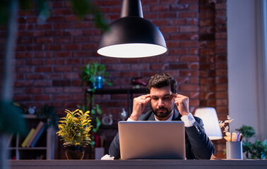 Indian businessman at desk holding head in pain, suffering from stress and workplace burnout