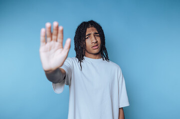 Young African man with curly hair shows hand gesture against blue background conveying a message of...