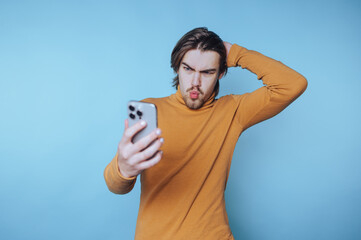 Young man in yellow sweater gazing at his smartphone with a pensive expression against a light blue...