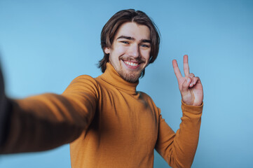 Young man taking a selfie while smiling and showing a peace sign against a light blue background
