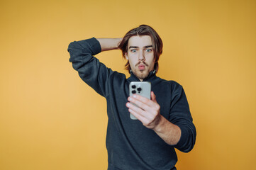 Young man checks his phone with surprise against a bright yellow backdrop while striking a casual...