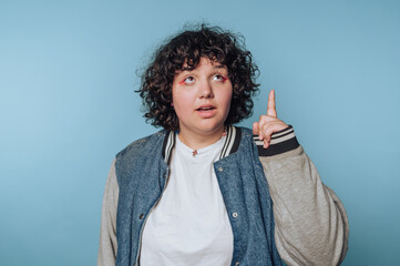 Young person with curly hair gesturing while thinking in front of a blue background in a casual...