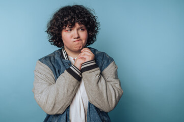 Child with curly hair looking thoughtful against a blue background while holding their hands...