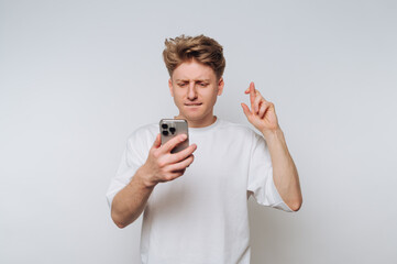Young man checks phone with a hopeful expression while crossing fingers for good luck in a bright,...