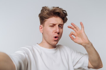 Young man making an okay gesture while taking a selfie in a simple indoor setting