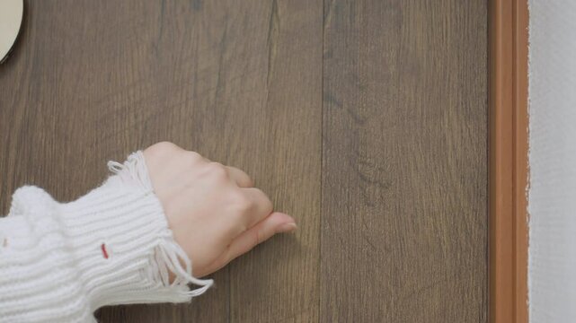 Close up of female hand in white knitted sweater knocking twice on wooden door in calm hallway as guide prepares to open door for session, showing gesture of arrival and request for entry
