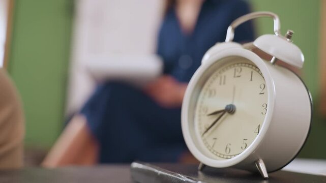 Classic white clock placed on table over notebook in foreground, with blurred view of specialist seated on sofa holding pen and writing note in calm contemplative room setting