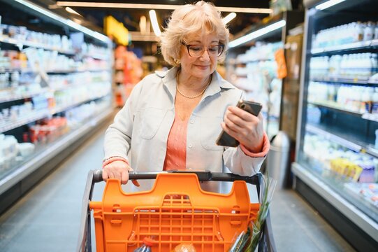 Happy senior woman looking at grocery store aisle with cellphone in hand - Powered by Adobe