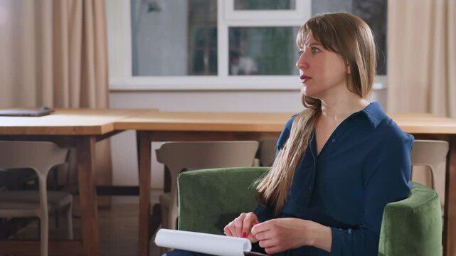 Cognitive coach speaking intentionally to female patient holding pen and notebook on lap in modern therapy room with wooden table chairs window and curtains in warm lighting setting