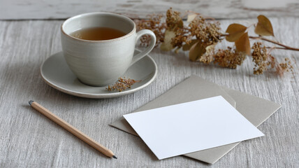 Cup of tea, blank greeting card with envelope, pencil and dry flowers create a cozy autumnal still life on a rustic table