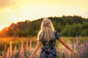 Blonde woman with long hair in a floral dress standing in a blooming wildflower field during golden sunset. Serene summer nature scene, peaceful countryside mood, romantic and natural lifestyle.