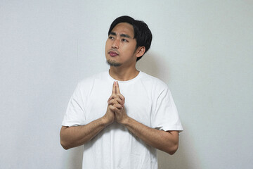 A young Asian man in a white t-shirt looks upward with his hands pressed together in a thoughtful...